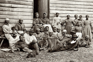 A historical black-and-white photo of a group of men, women, and children posed outside a wooden building.