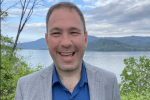 A man in a gray suit smiles by a lake, surrounded by trees and mountains under a cloudy sky.