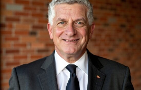 A man in a suit smiles in front of a brick wall background.