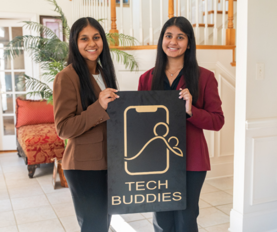Two young women in blazers holding a sign that reads "Tech Buddies" in an indoor setting.