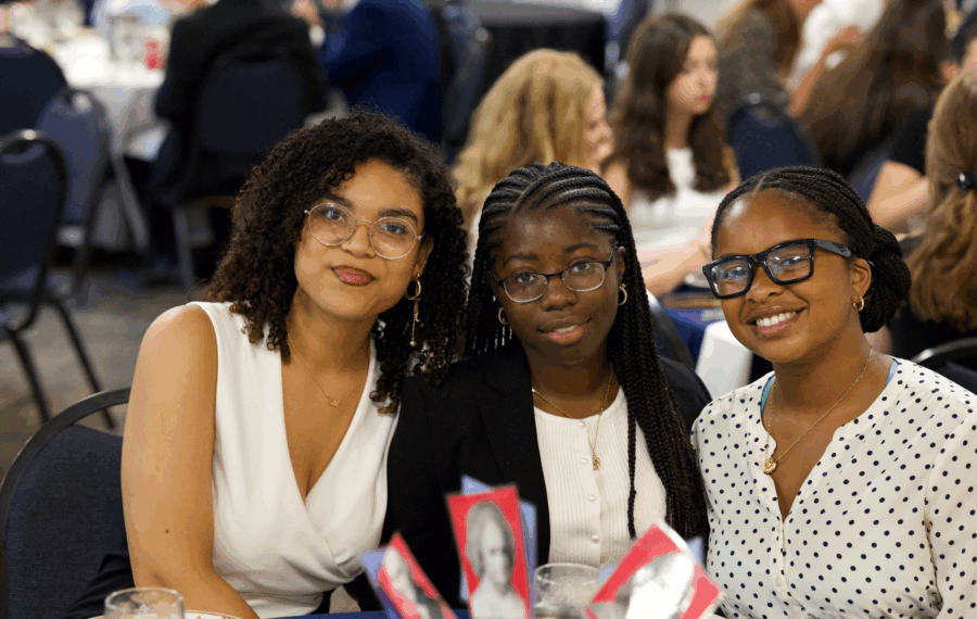 Three women sitting together at a dining table, smiling for the camera, in a formal setting with people in the background.