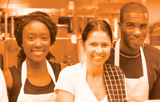 A group of five diverse people in a kitchen, smiling and wearing aprons, posing together.