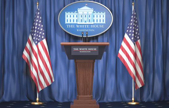 Podium with "The White House" seal, flanked by American flags, set against a blue curtain backdrop.