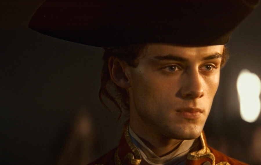 Young man in a historical costume with a large hat, looking serious and focused against a dimly lit background.