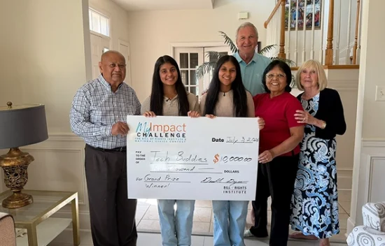 Two young women hold a large check for $10,000 with family members in a home setting.