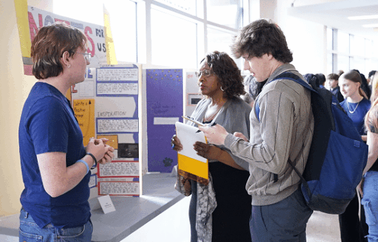 Three individuals engage in discussion at an informational event, with displays and posters in the background.